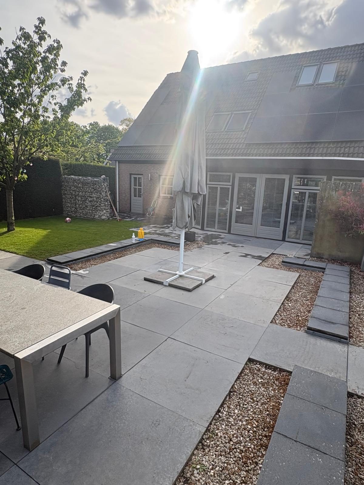 Terras met grote grijze tegels, grindranden, een tuintafel en gazon achter een woonhuis in de zon