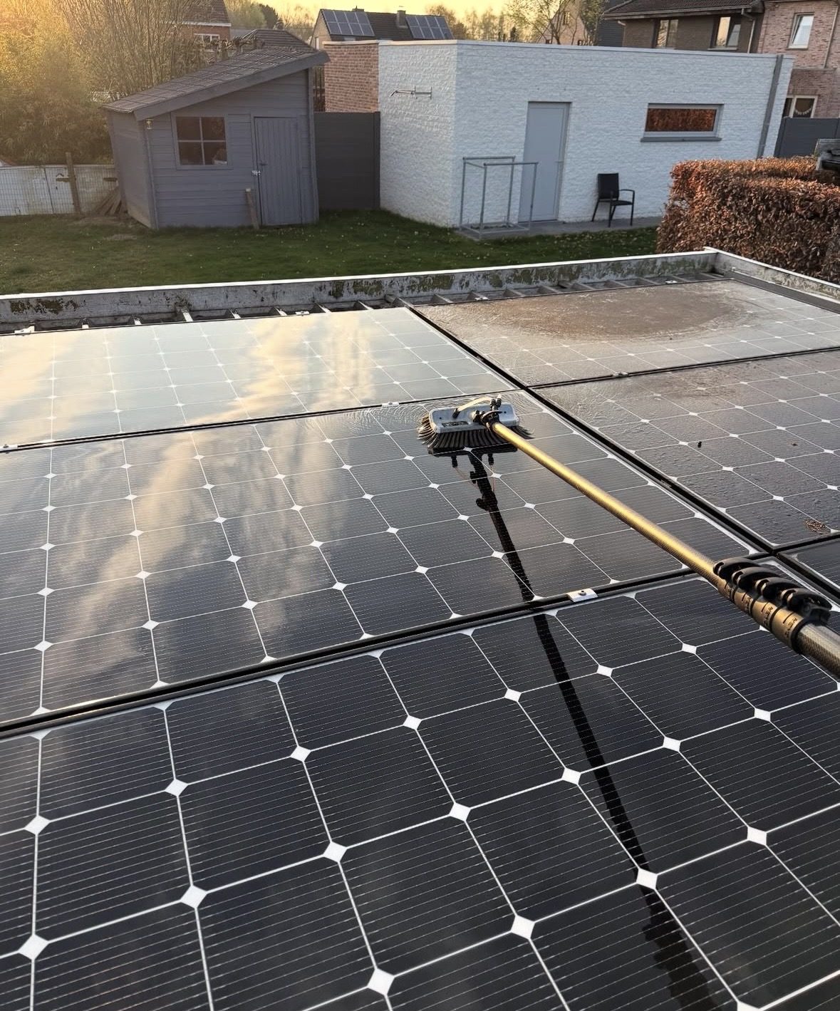 Zonnepanelen op een plat dak worden gereinigd met een borstel aan een telescoopsteel: de linker panelen glanzen al, de rechter zijn nog dof en vuil